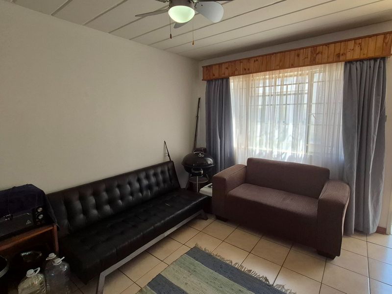 A living room featuring tiled flooring, a ceiling fan, and a large window with a wooden pelmet, offering natural light and access to an outdoor area.