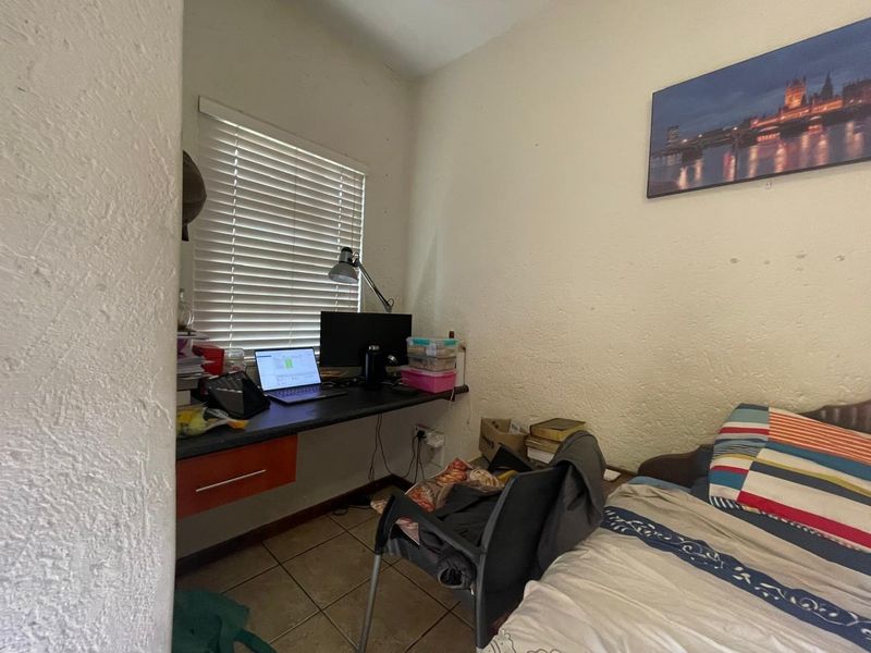 A compact bedroom featuring a built-in desk, tiled flooring, and venetian blinds, offering a functional space that could benefit from cosmetic updates.