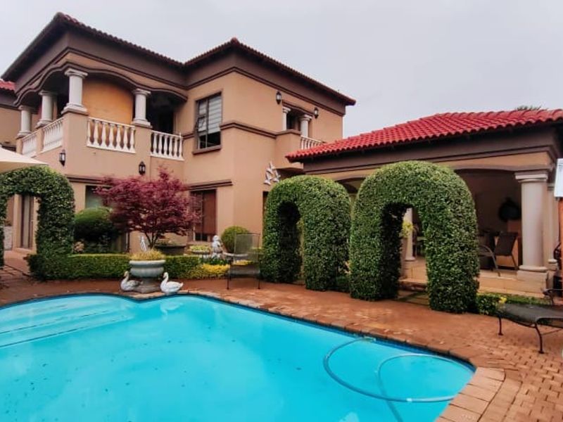 A well-maintained outdoor pool area featuring brick paving, a covered patio, and a multi-story residence with a balcony and tiled roof.