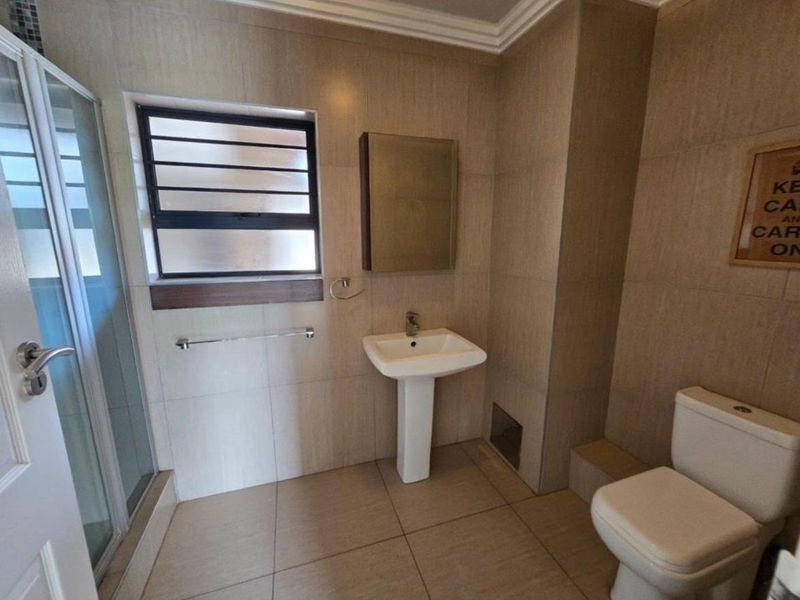 A functional bathroom featuring a pedestal basin, shower enclosure, toilet, and neutral wall and floor tiling with a louvered window for ventilation.