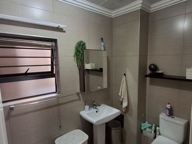 A functional bathroom featuring a pedestal washbasin, toilet, and neutral wall tiling. The space includes a medicine cabinet and window with security bars.