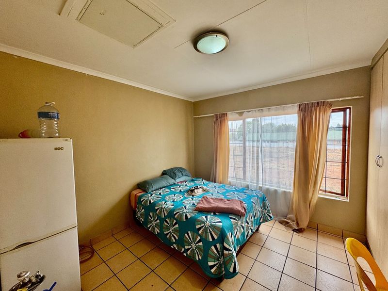 A bedroom featuring tiled flooring, built-in cupboards, and a large window, offering a functional space in need of cosmetic updates.