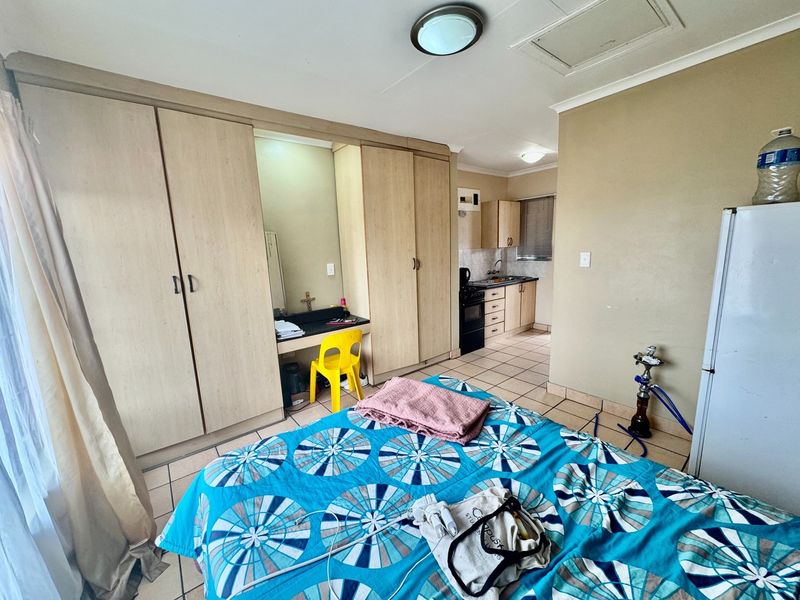 Open-plan studio apartment featuring built-in bedroom cupboards with integrated vanity, tiled flooring throughout, and a compact functional kitchen area.