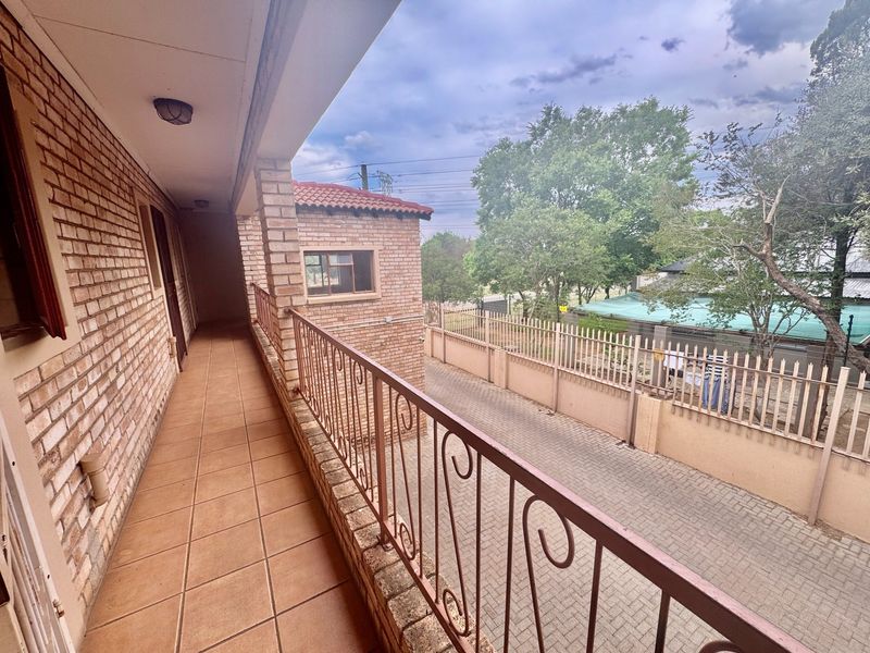 A covered balcony featuring tiled flooring, brick walls, and a wrought iron railing overlooking the paved driveway area.