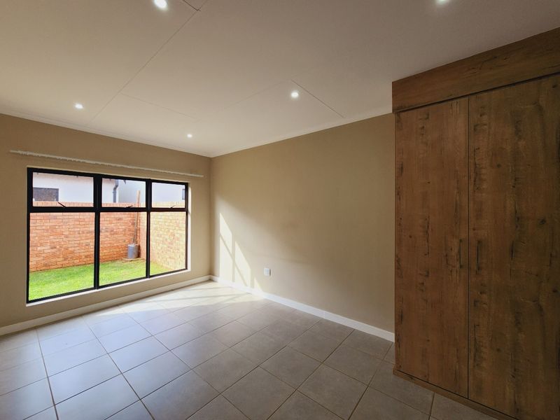 A bright, empty bedroom featuring neutral tiled flooring, recessed downlighting, and a modern built-in wooden wardrobe with views of the rear garden.