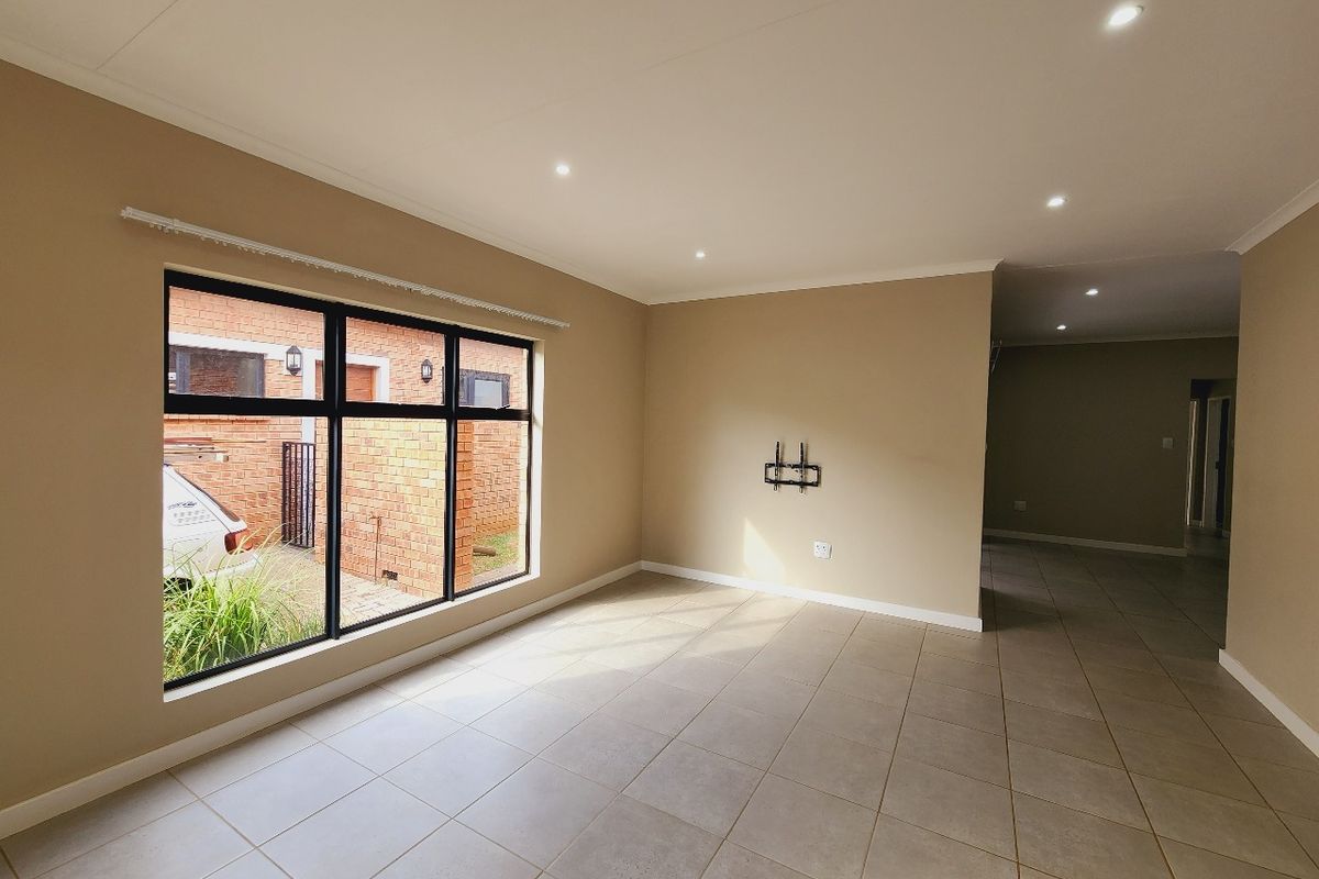 A bright, open-plan living area featuring neutral wall tones, large aluminium-framed windows, and modern tiled flooring with recessed downlighting.