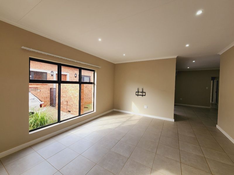 A bright, open-plan living area featuring neutral wall tones, large aluminium-framed windows, and modern tiled flooring with recessed downlighting.