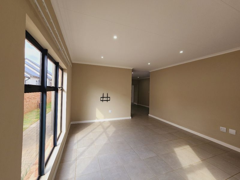 A bright, open-plan living area featuring neutral wall tones, large windows, tiled flooring, and recessed downlighting, providing a clean and versatile space.