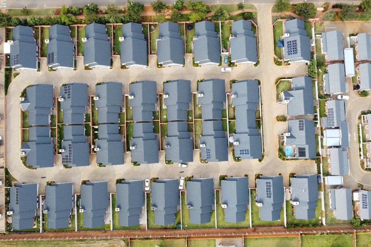 An aerial view of a residential townhouse complex featuring uniform roof structures, paved access roads, and private garden plots.