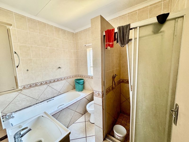 A dated bathroom featuring a built-in bathtub, separate shower enclosure with a damaged door, toilet, and vanity basin with neutral ceramic tiling.