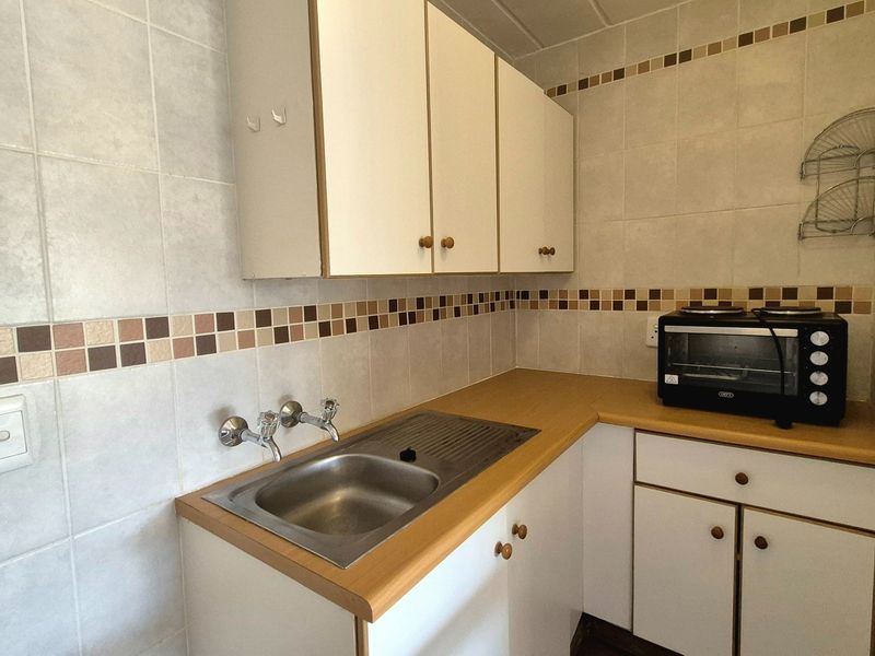 A compact kitchen area featuring dated white cabinetry, a laminate countertop, stainless steel sink, and tiled backsplash with decorative accents.