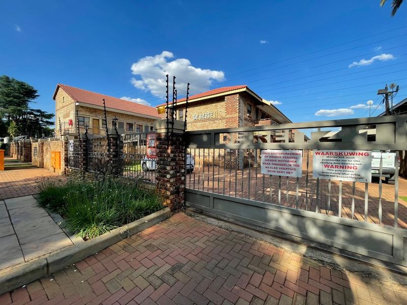 Front entrance view of the apartment complex featuring a secure metal gate, brick boundary wall, electric fencing, and paved driveway access.