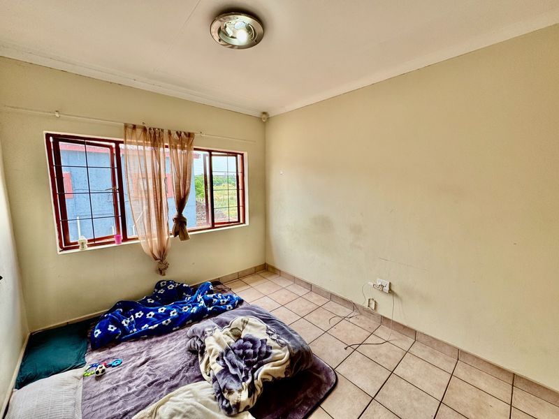 A simple bedroom featuring tiled flooring, a ceiling light, and a window with security bars, currently requiring minor wall repairs and fresh paint.