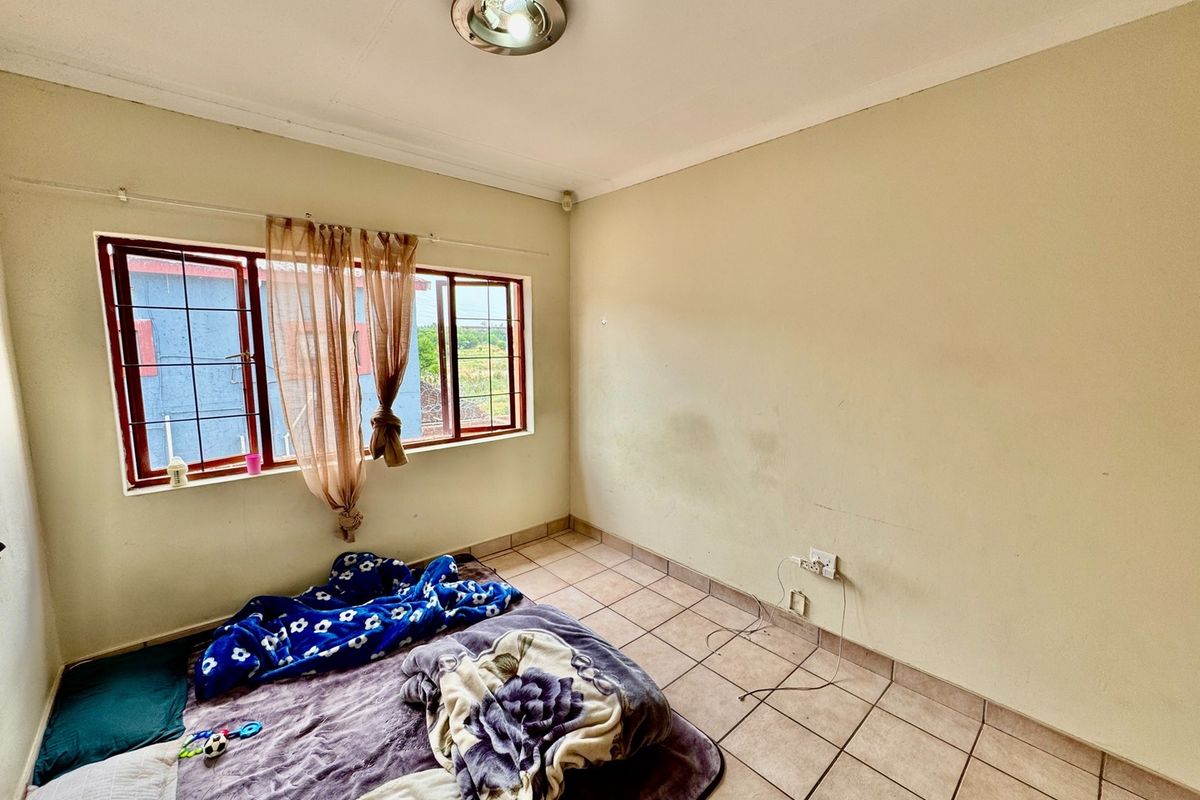 A simple bedroom featuring tiled flooring, a ceiling light, and a window with security bars, currently requiring minor wall repairs and fresh paint.