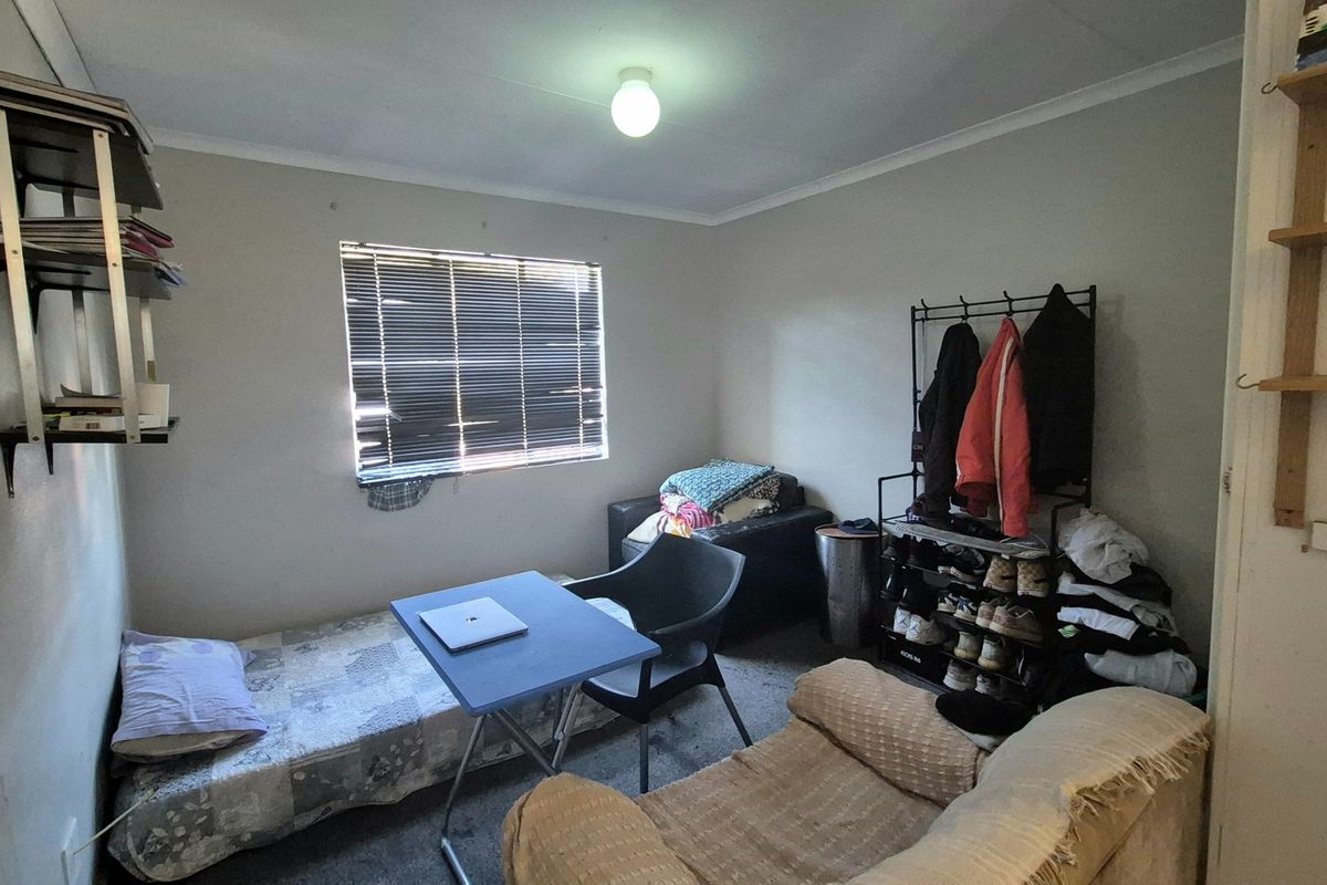 A compact bedroom featuring a window with venetian blinds, wall-mounted shelving, and a standard ceiling light fixture.