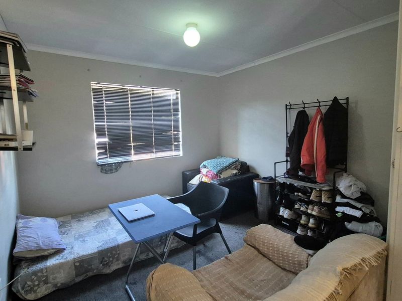 A compact bedroom featuring a window with venetian blinds, wall-mounted shelving, and a standard ceiling light fixture.