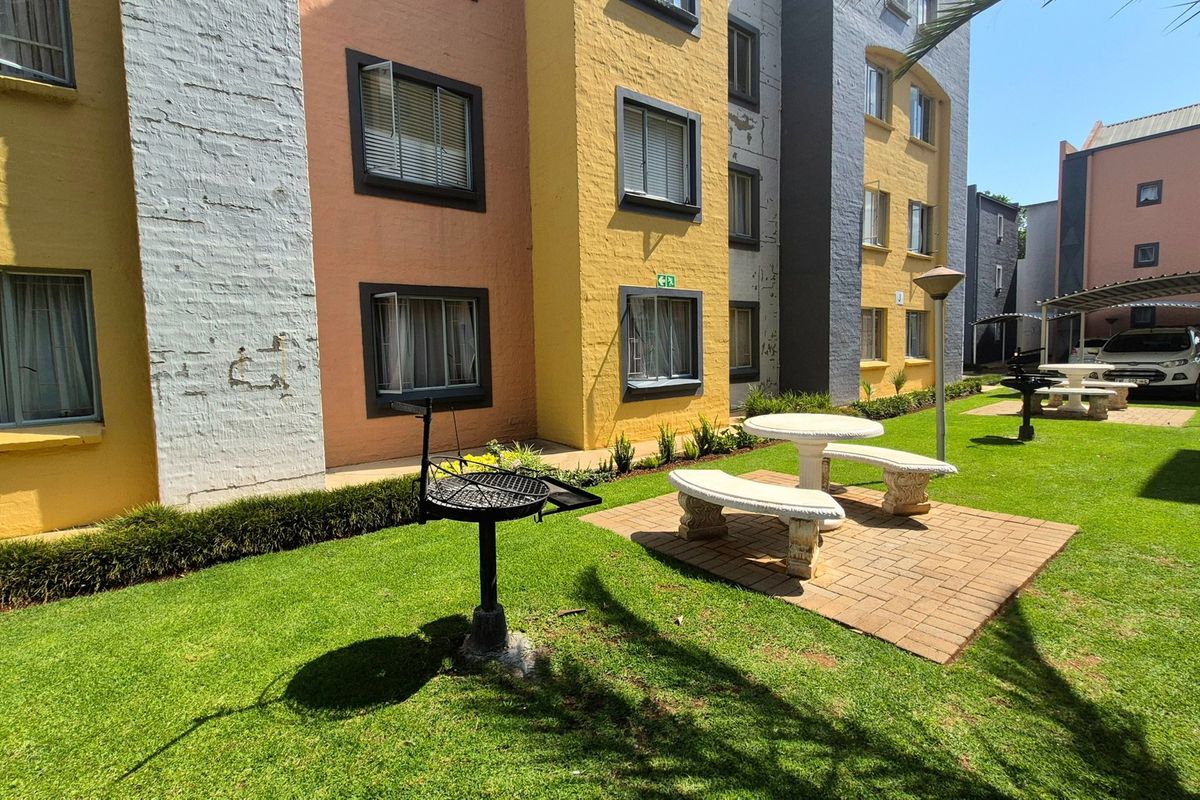 Communal garden area featuring a paved patio, fixed stone seating, and a built-in braai stand set against the apartment building exterior.