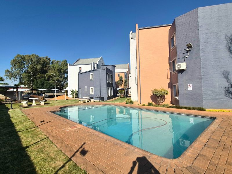Communal swimming pool area featuring brick paving, landscaped lawn, and multi-story apartment building facades.
