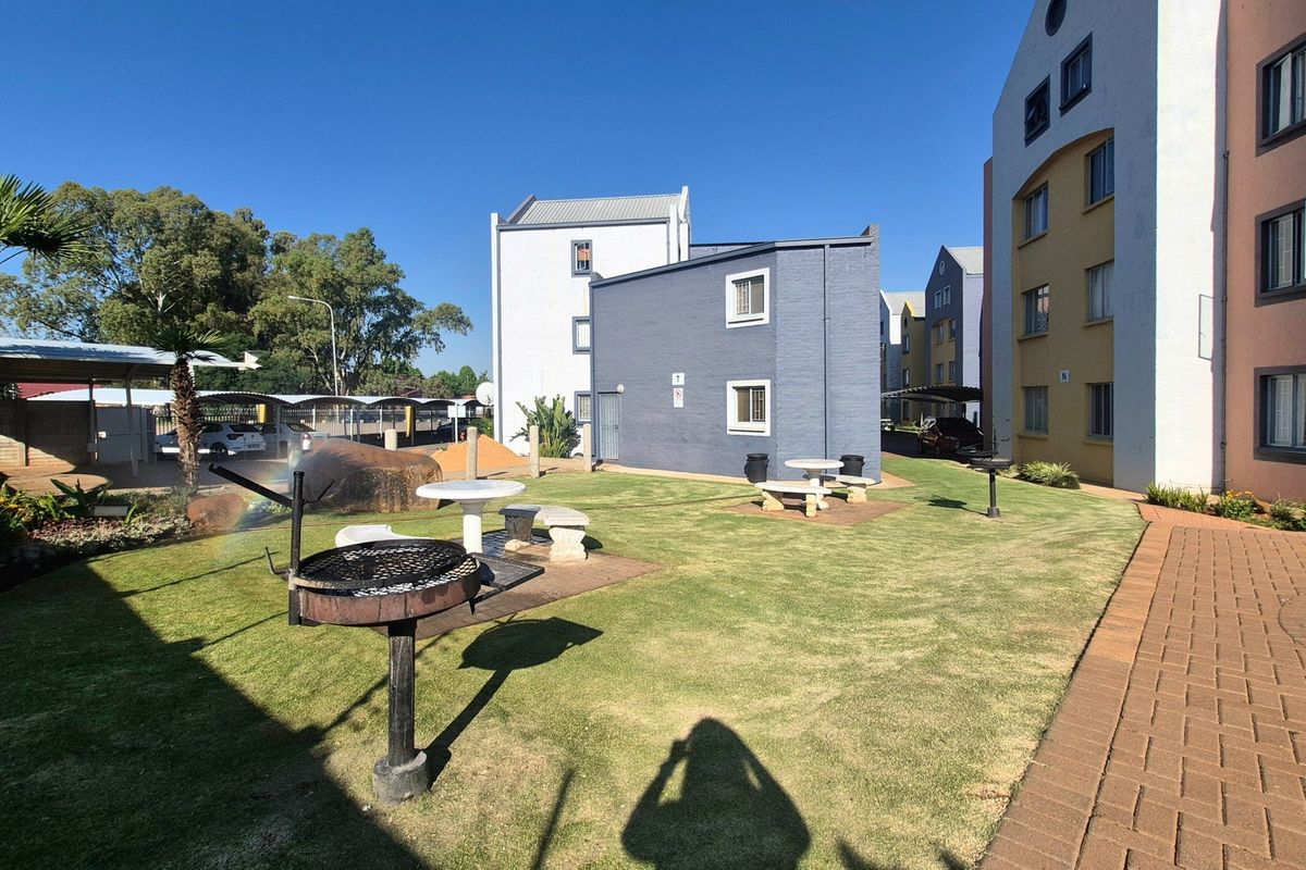 A well-maintained communal garden area featuring a lawn, paved pathways, built-in braai facilities, and stone seating, situated between apartment blocks.