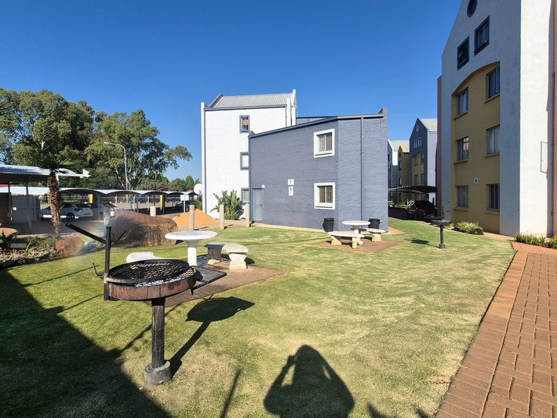 A well-maintained communal garden area featuring a lawn, paved pathways, built-in braai facilities, and stone seating, situated between apartment blocks.