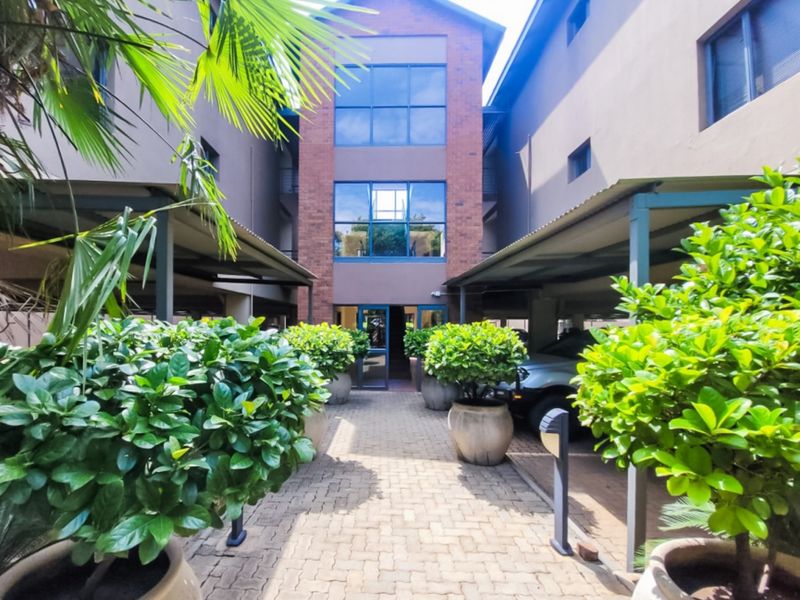 Front entrance view of the apartment complex featuring a brick facade, glass windows, and covered parking bays with paved access.