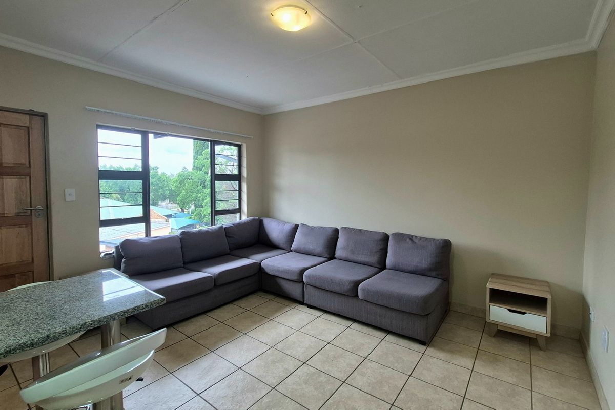 A living room area featuring neutral tiled flooring, a large window providing natural light, and a coved ceiling with a central light fixture.