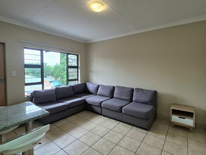 A living room area featuring neutral tiled flooring, a large window providing natural light, and a coved ceiling with a central light fixture.