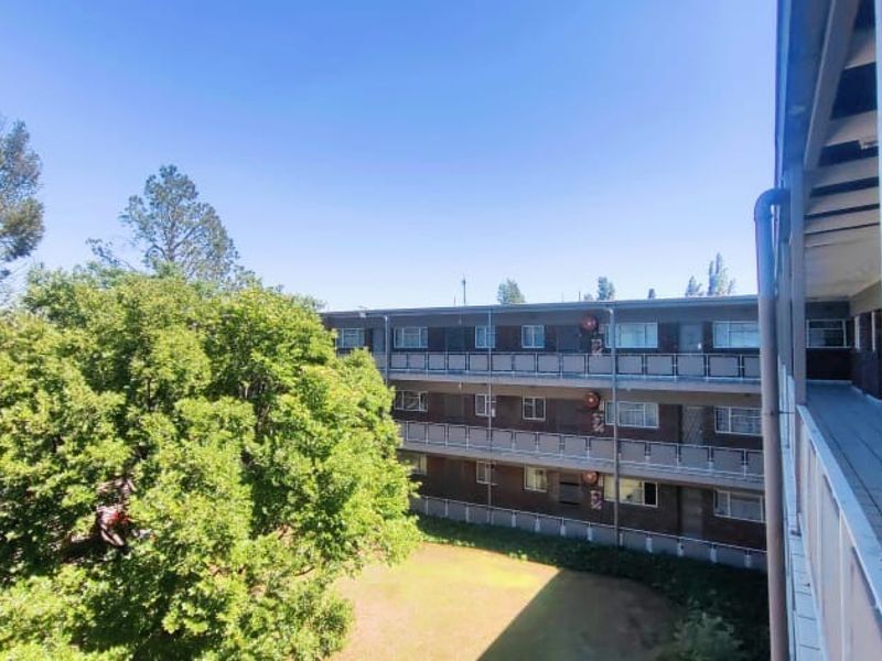 View of the apartment complex exterior, featuring a covered balcony with metal railings overlooking the communal garden and adjacent building facade.