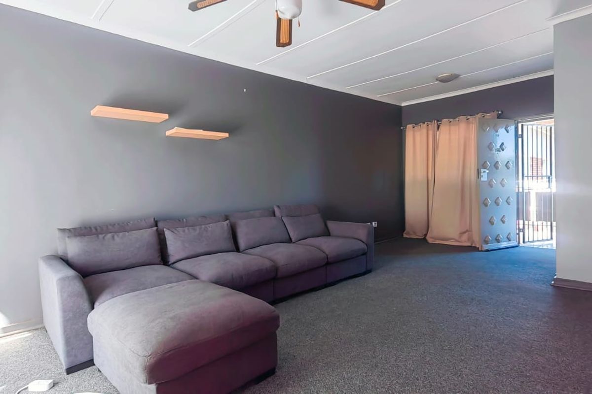 A living room featuring grey walls, carpeted flooring, a ceiling fan, and a secure metal entrance gate.