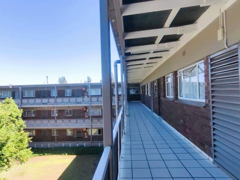 An exterior walkway balcony featuring tiled flooring, brick wall finishes, and security gates, overlooking the apartment complex grounds.