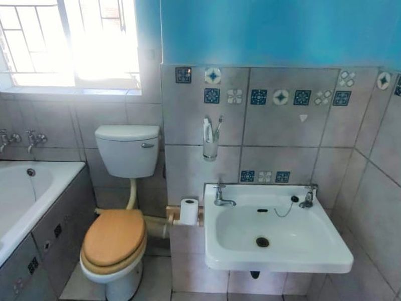 A functional bathroom featuring a bathtub, wall-mounted basin, and toilet, finished with dated decorative wall tiles and security bars on the window.