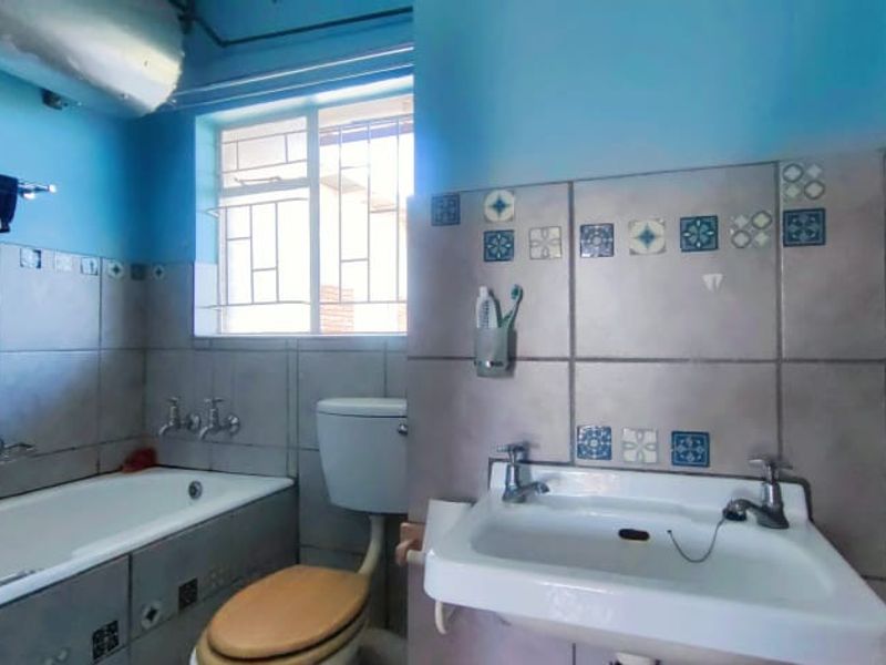 A dated bathroom featuring a bathtub, pedestal basin, and toilet, with decorative tiled walls and security bars on the window.