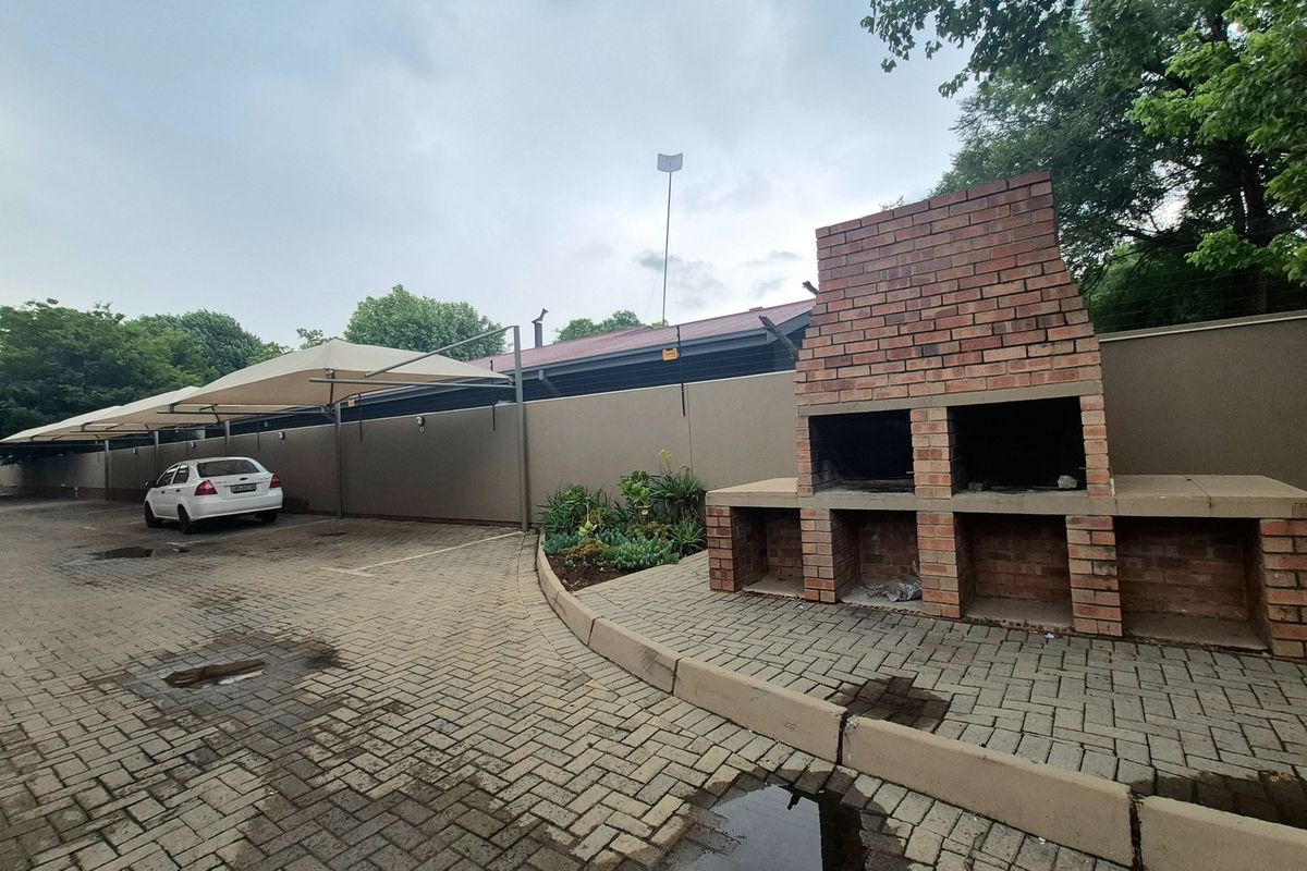 Outdoor communal area featuring a brick braai, paved parking area with shade cloth carports, and a perimeter boundary wall.