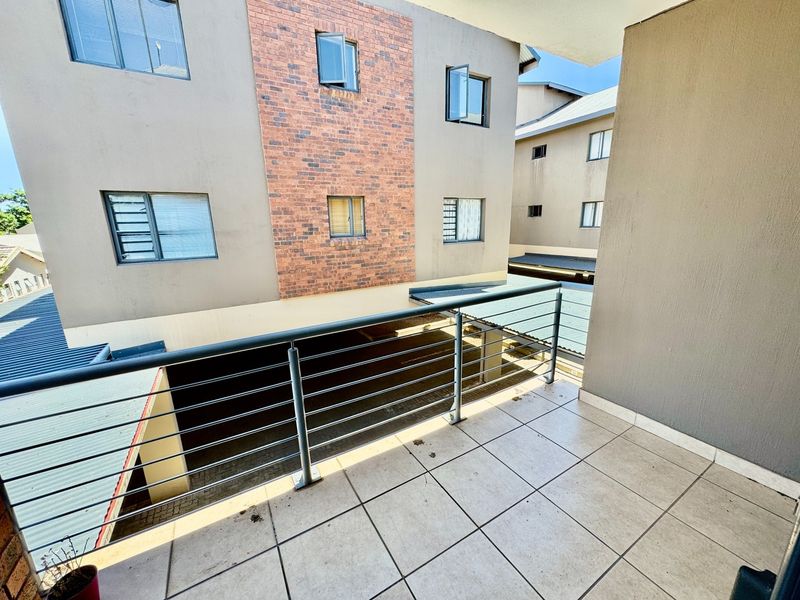 A covered balcony featuring tiled flooring and a metal balustrade, overlooking the apartment complex courtyard with brick and rendered facade details.