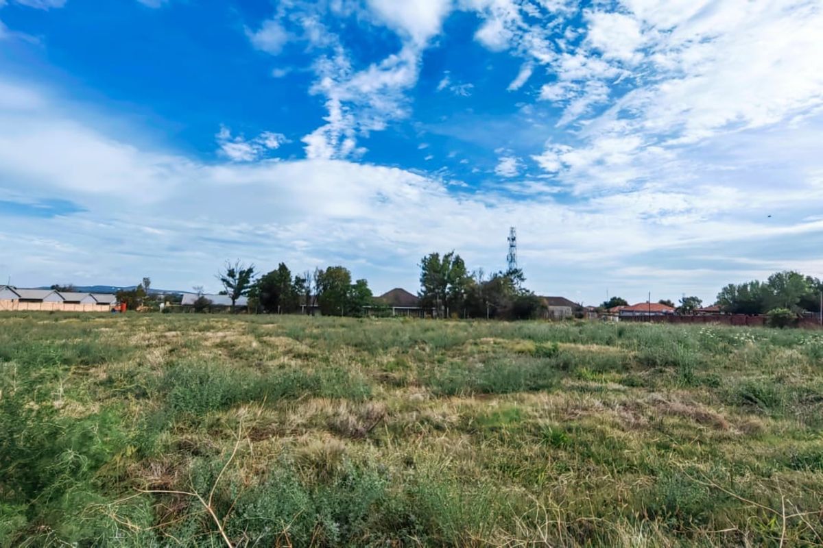 A wide-angle view of a vacant residential plot featuring open grassland with surrounding neighborhood properties in the distance.