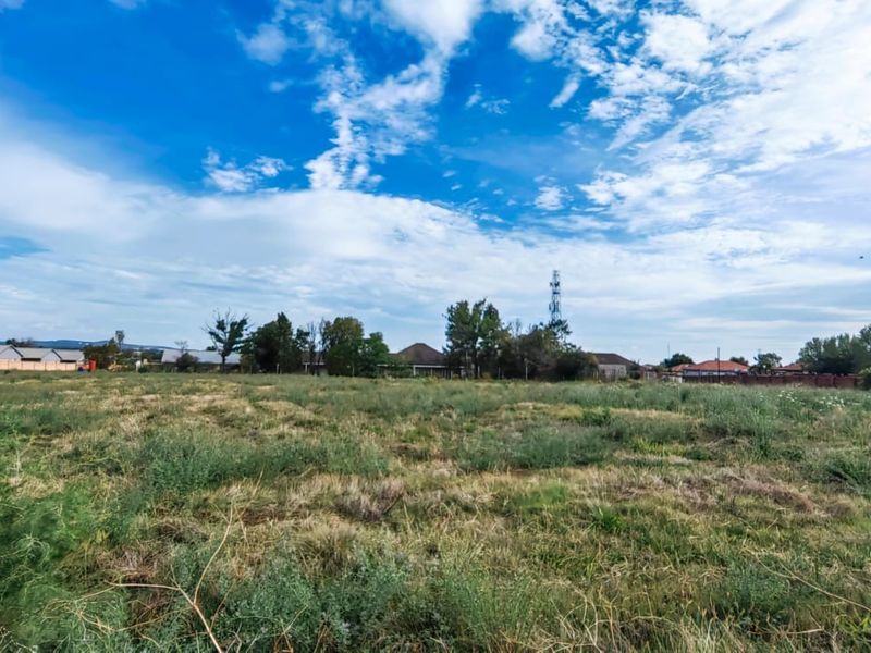 A wide-angle view of a vacant residential plot featuring open grassland with surrounding neighborhood properties in the distance.