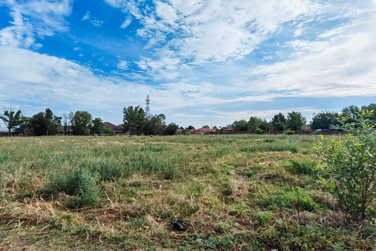 A wide-angle view of a vacant residential plot featuring open grassland with surrounding neighborhood trees and infrastructure in the distance.