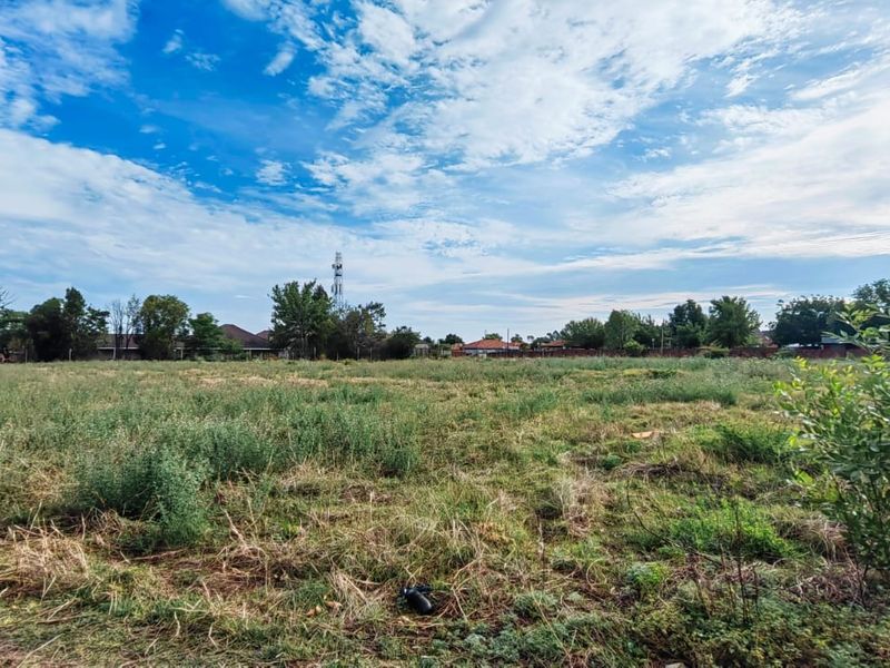 A wide-angle view of a vacant residential plot featuring open grassland with surrounding neighborhood trees and infrastructure in the distance.