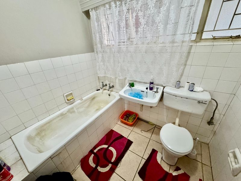 A dated bathroom featuring a standard bathtub, wall-mounted basin, and toilet, with ceramic tiling throughout and a window with security bars.
