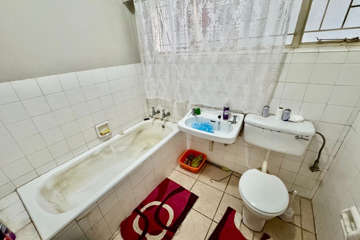 A dated bathroom featuring a standard bathtub, wall-mounted basin, and toilet, with ceramic tiling throughout and a window with security bars.