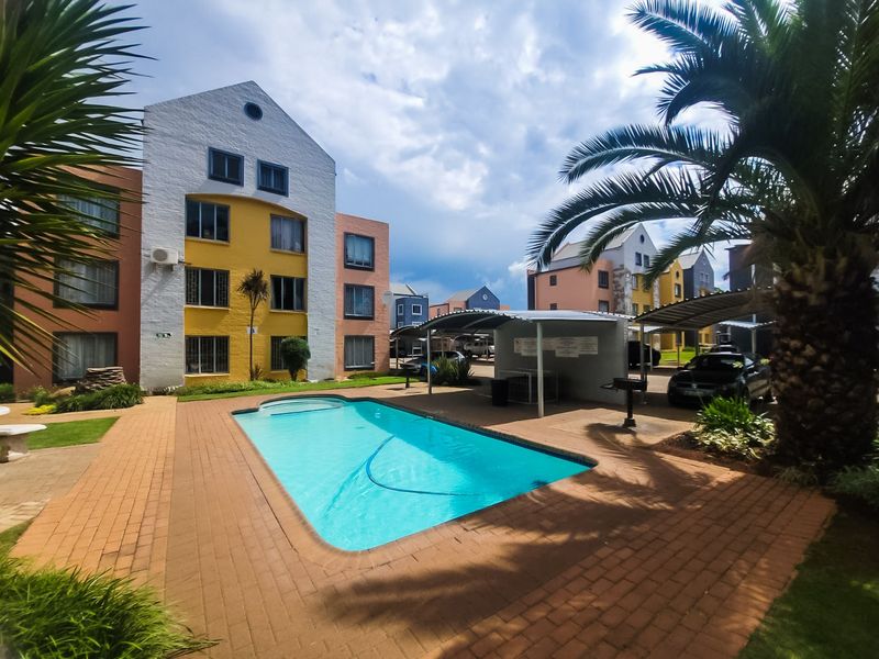 A well-maintained communal pool area featuring a paved deck, surrounding landscaped gardens, and covered parking bays within the apartment complex.