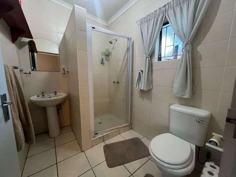 A functional bathroom featuring a pedestal basin, a glass-enclosed shower, and a standard toilet with neutral wall and floor tiling.