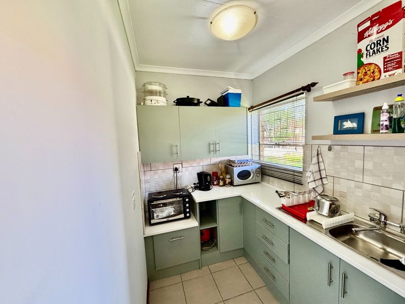 Compact kitchen featuring sage green cabinetry, laminate countertops, tiled backsplash, stainless steel sink, and functional storage space with floating shelves.