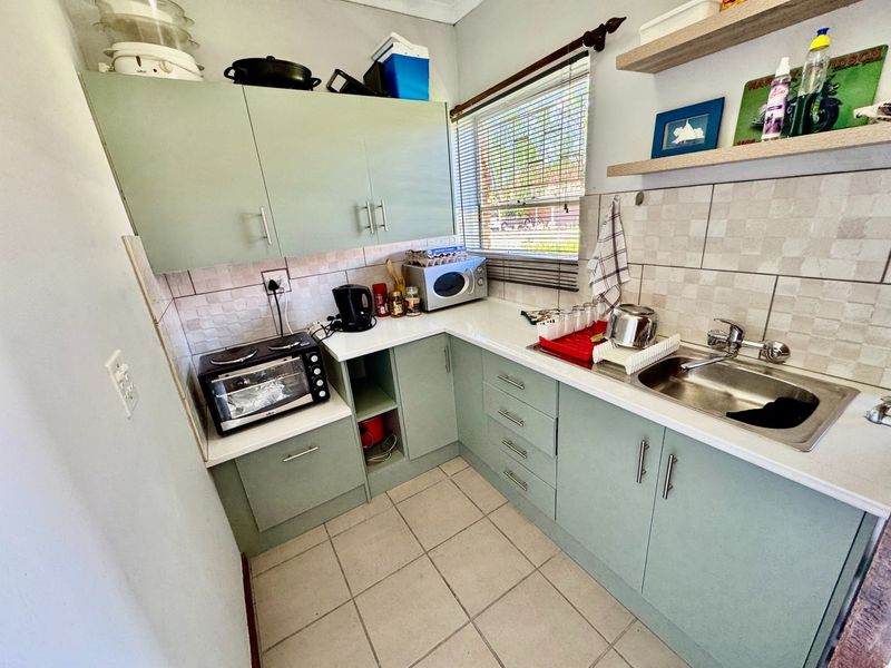 Compact kitchen featuring sage green cabinetry, white countertops, tiled splashback, stainless steel sink, and floating wall shelves.