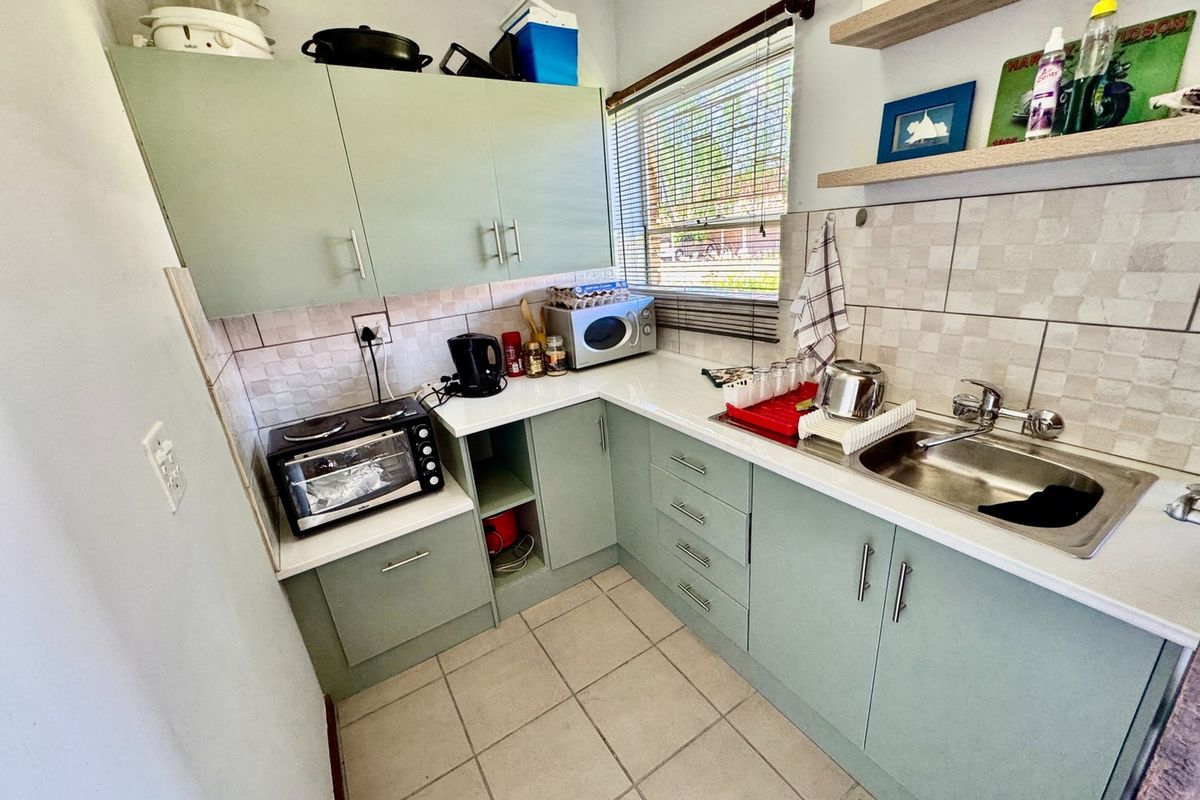 Compact kitchen featuring sage green cabinetry, white countertops, tiled splashback, stainless steel sink, and floating wall shelves.