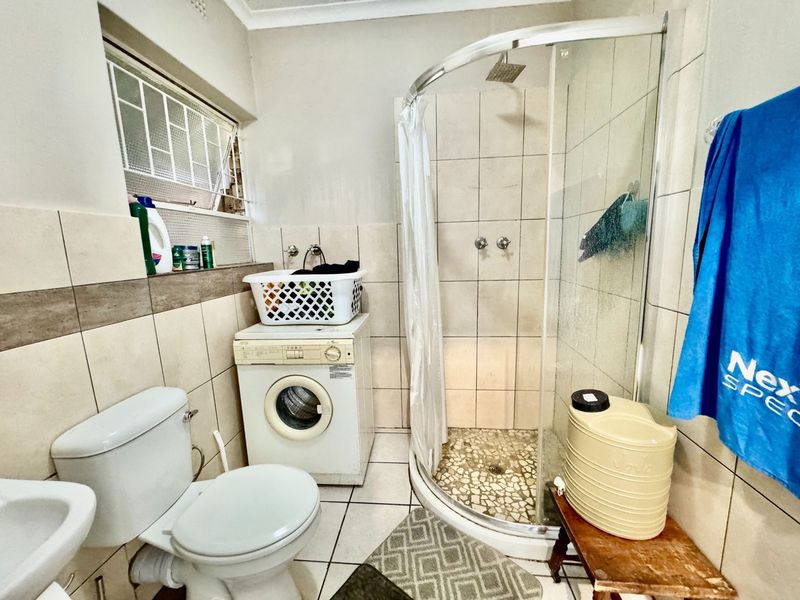 A functional bathroom featuring a corner shower enclosure, toilet, wash basin, and integrated laundry space with tiled finishes throughout.