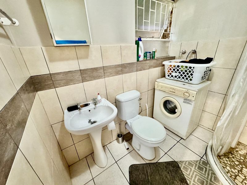Compact bathroom featuring a pedestal basin, toilet, shower enclosure, and integrated laundry plumbing, finished with neutral wall and floor tiling.