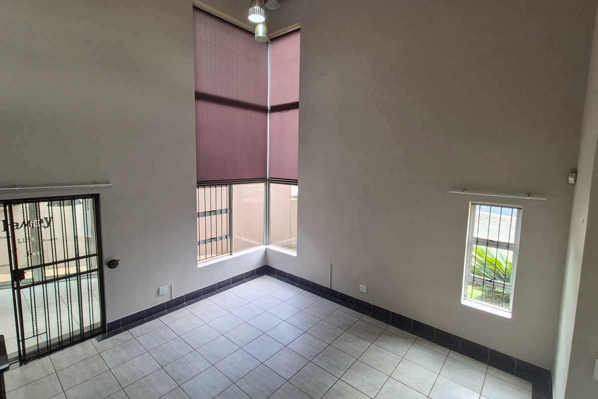 An empty living area featuring high ceilings, tiled flooring, a corner window with roller blinds, and a secure metal entrance gate.
