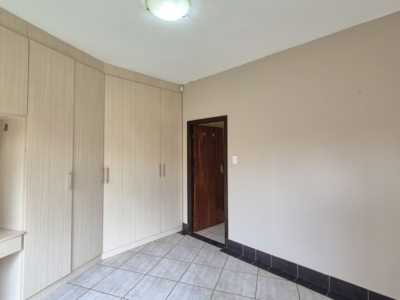A bright, empty bedroom featuring light wood-toned built-in cupboards with an integrated vanity and neutral tiled flooring.