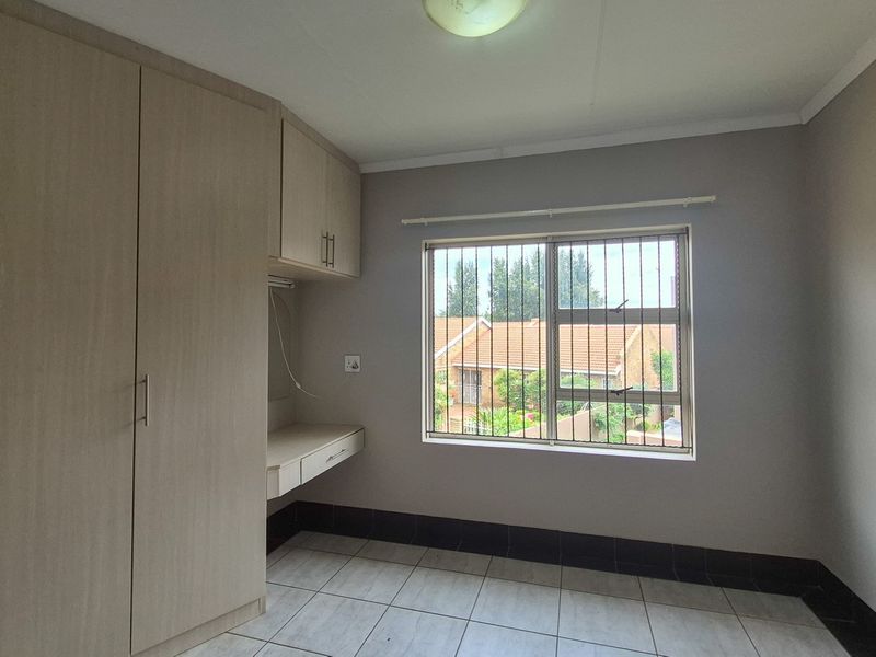 A clean, empty bedroom featuring neutral tiled flooring, built-in cupboards, a integrated desk area, and a window fitted with security bars.